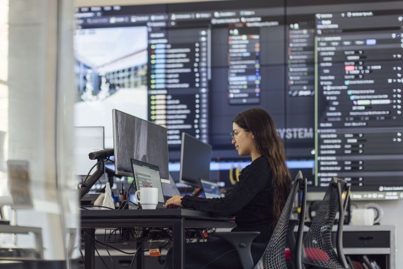 Female student with glasses and long dark hair sitting in front of a computer, with large screens displaying complex computing in the background.