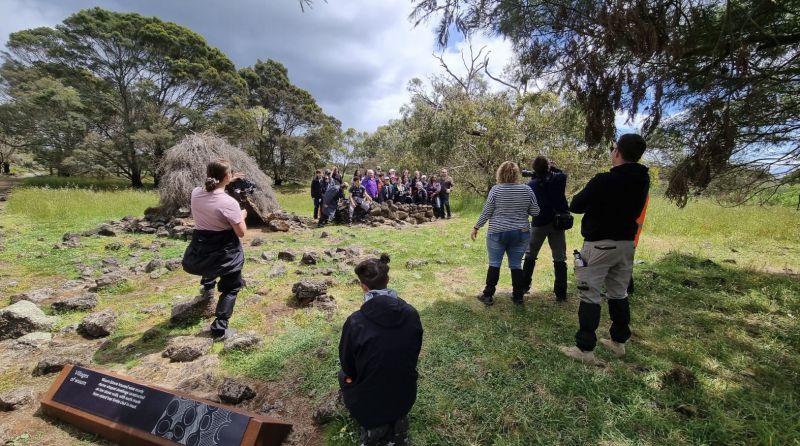 A group of people stand and watch Elders and students posing for camera out in a grassy and tree-filled area of Budj Bim. 