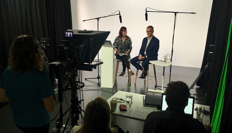 A man and a woman sit on chairs in front of a camera crew in a studio, answering questions.