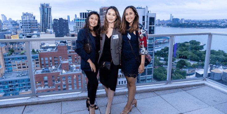 A group of three alumni smiling to camera on a balcony overlooking New York at an association event.