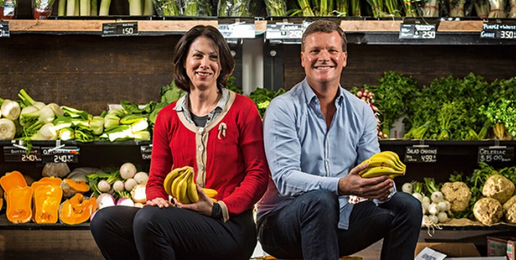 A man and woman smiling to camera holding a bunch of bananas with other produce in the background.