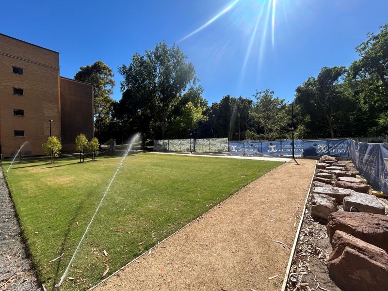 Grassy area with a path and irrigation sprinklers beside trees and a building.