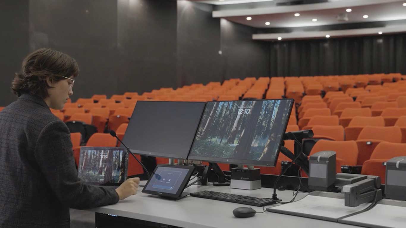 A woman adjusts the controls of her lectern in an empty lecture theatre.