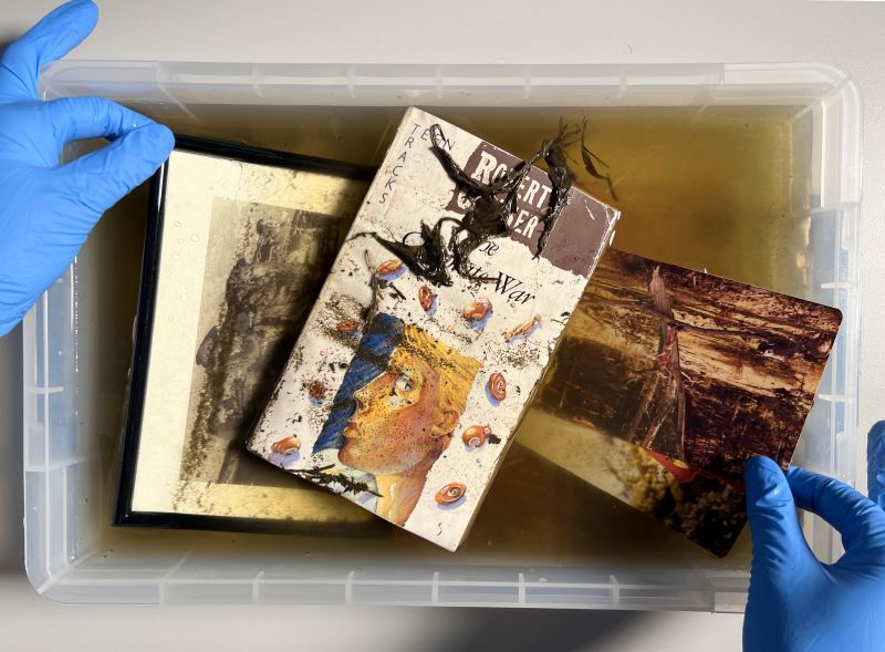 Two hands, wearing blue gloves, hold flood damaged items including a book and a framed picture partially submerged in a container of dirty water.
