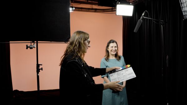 A woman with dark hair in a blue dress stands with her arms open and happy expression in front of an orange background in a studio setting. Another woman with dark hair in a black jacket snaps a clapperboard in front of her for her take..