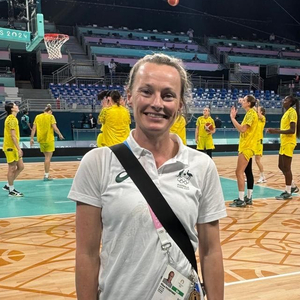 A female alum smiling to camera with basketball players playing on a court in the background.
                                class=