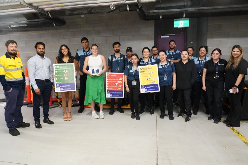 A group of around 20 people standing in an indoor waste facility and holding colourful waste education signage