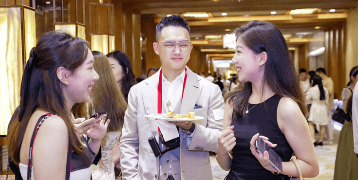 Three young Chinese alumni mingling at an event. One man is holding a plate of food.
