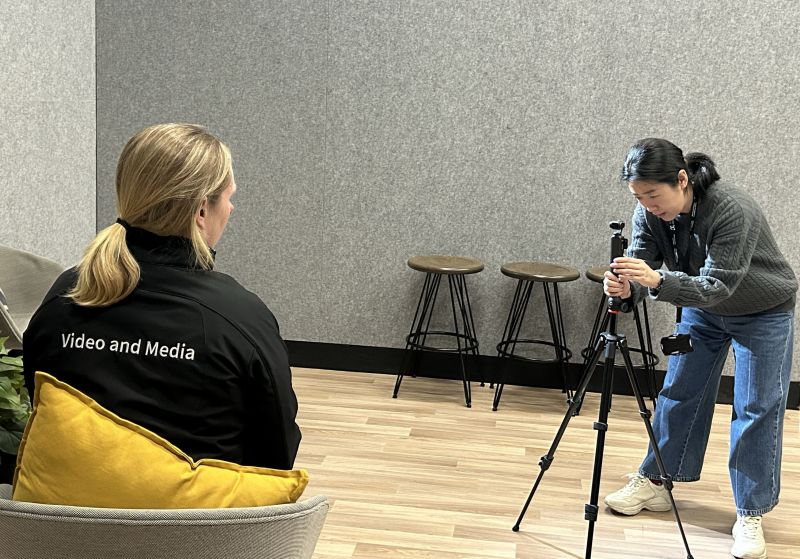 A woman adjusts her camera in front of a woman wearing a jacket that says 'Video and Media' on the back.
