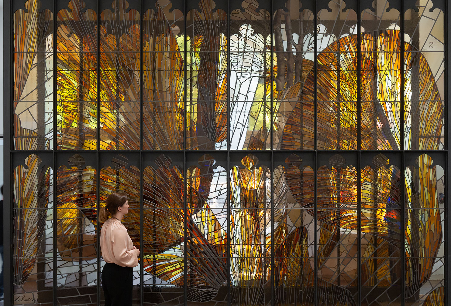 A woman stands in front of a large stained glass wall in yellow and brown