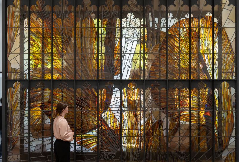 A woman stands in front of a large stained glass wall in yellow and brown