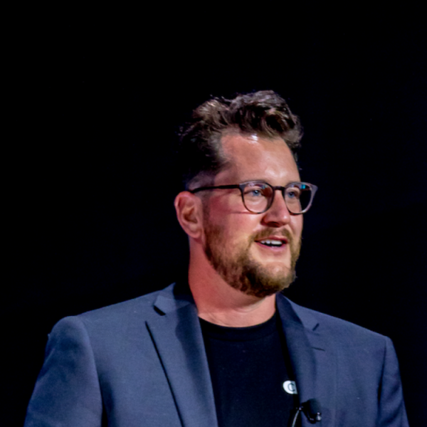 Person wearing glasses and a jacket standing against a dark background, speaking while facing to the side