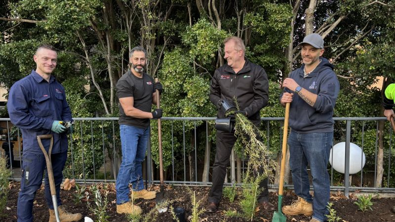 Four people standing outdoors in a garden bed holding shovels, with newly planted vegetation and trees in the background