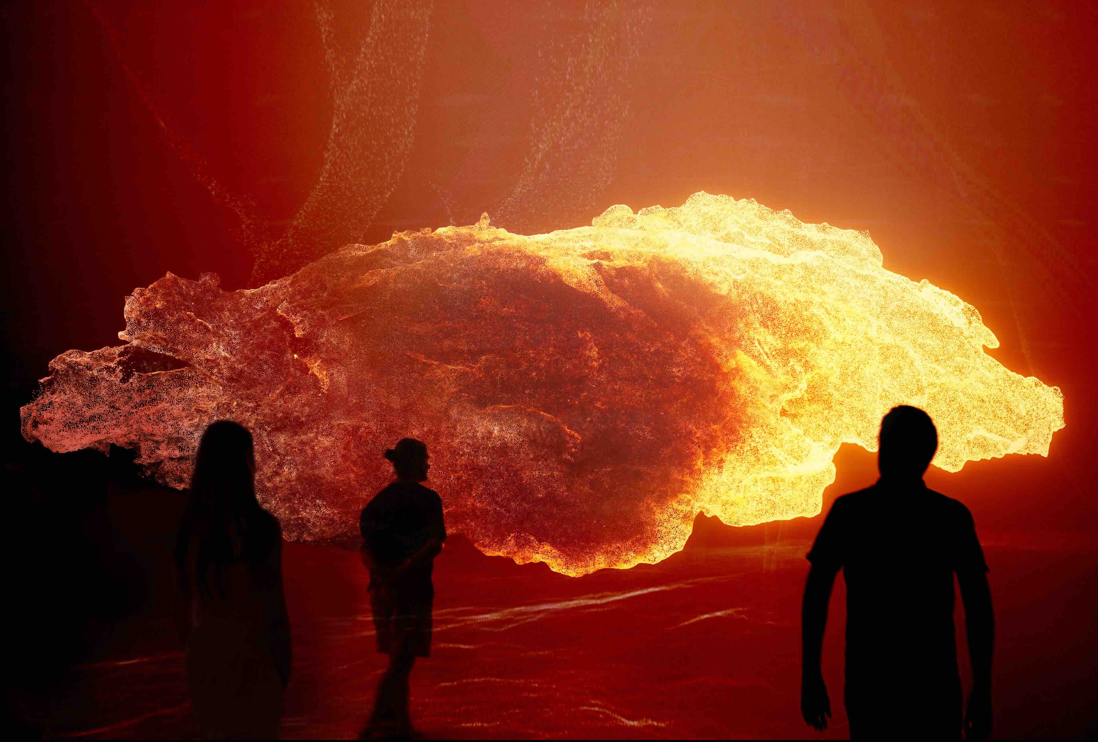 Large fiery installation or projection with silhouettes of people standing in front of it in a dark indoor space