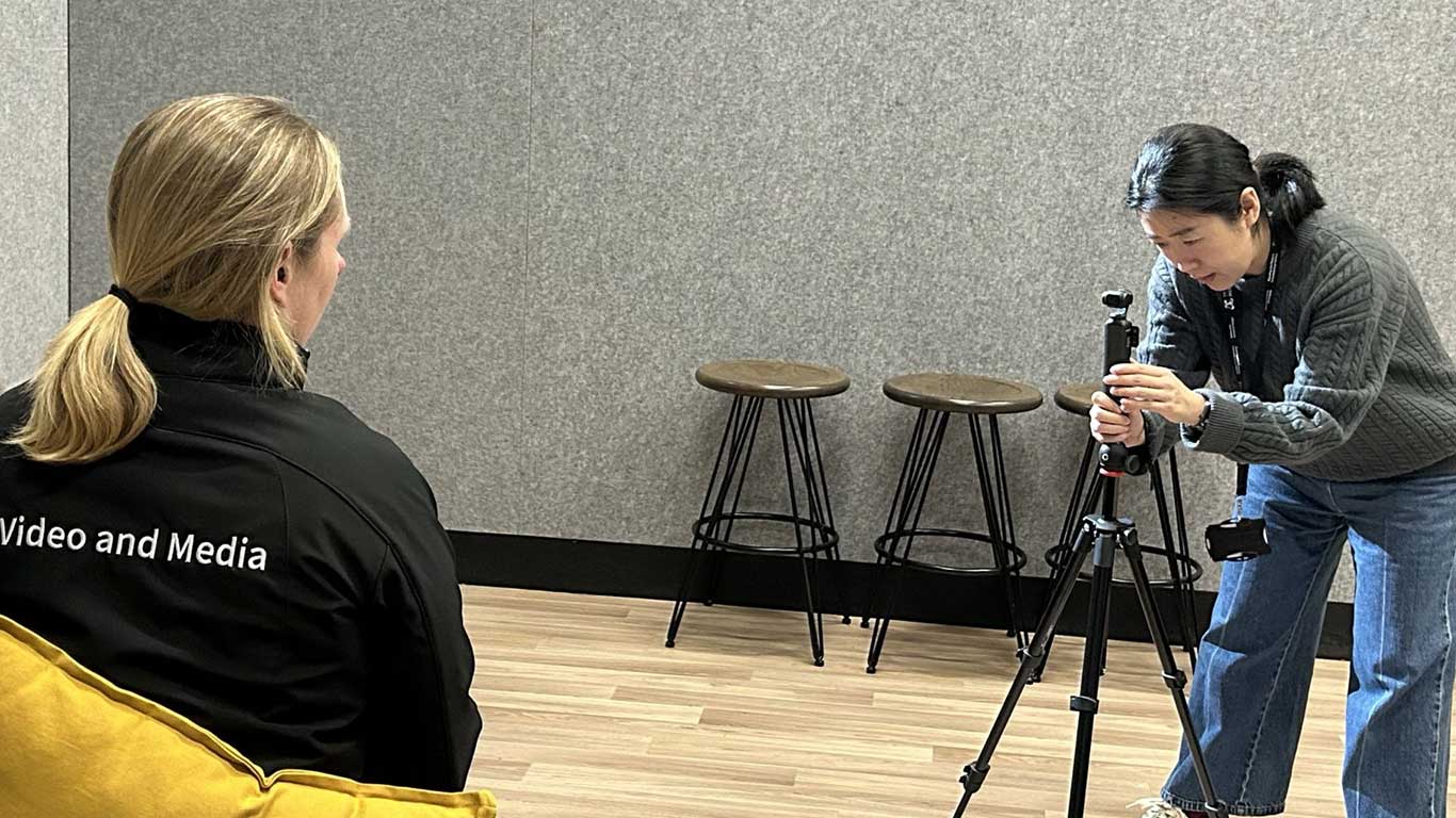 A woman adjusts a camera on a tripod as her Video and Media coach looks on.