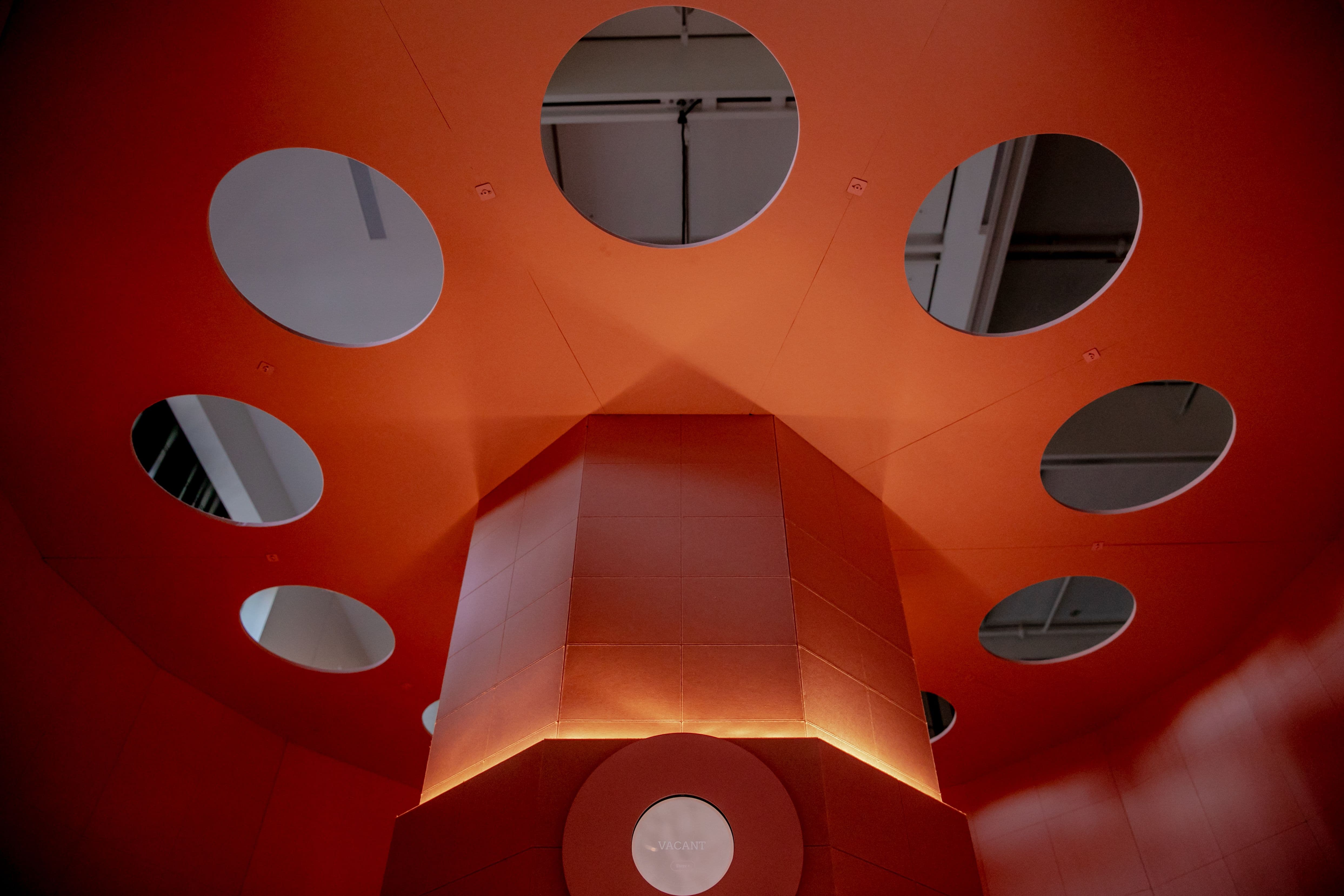 Ceiling of pink toilet cubicle, with circle cutouts through which the ceiling of the gallery can be seen