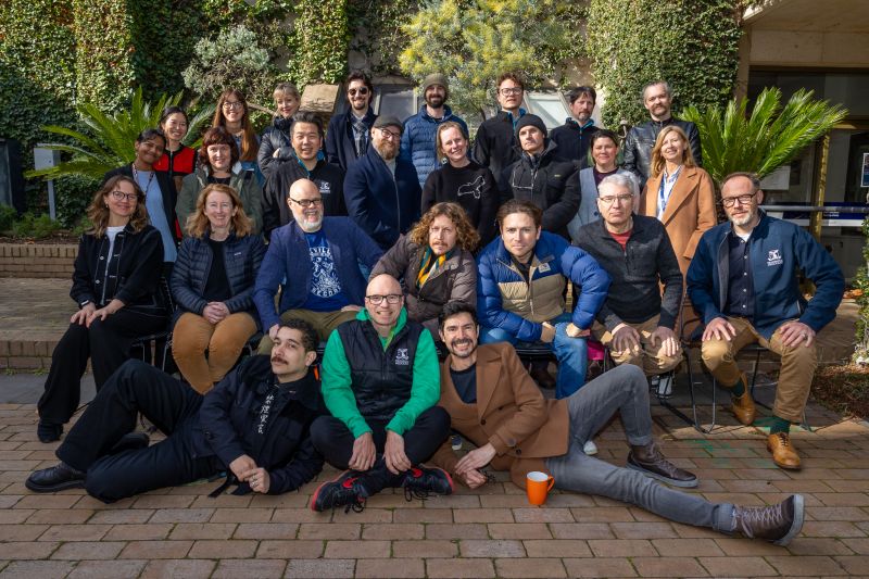 A group photo of a team of about 26 people with green plants and a building in the background.