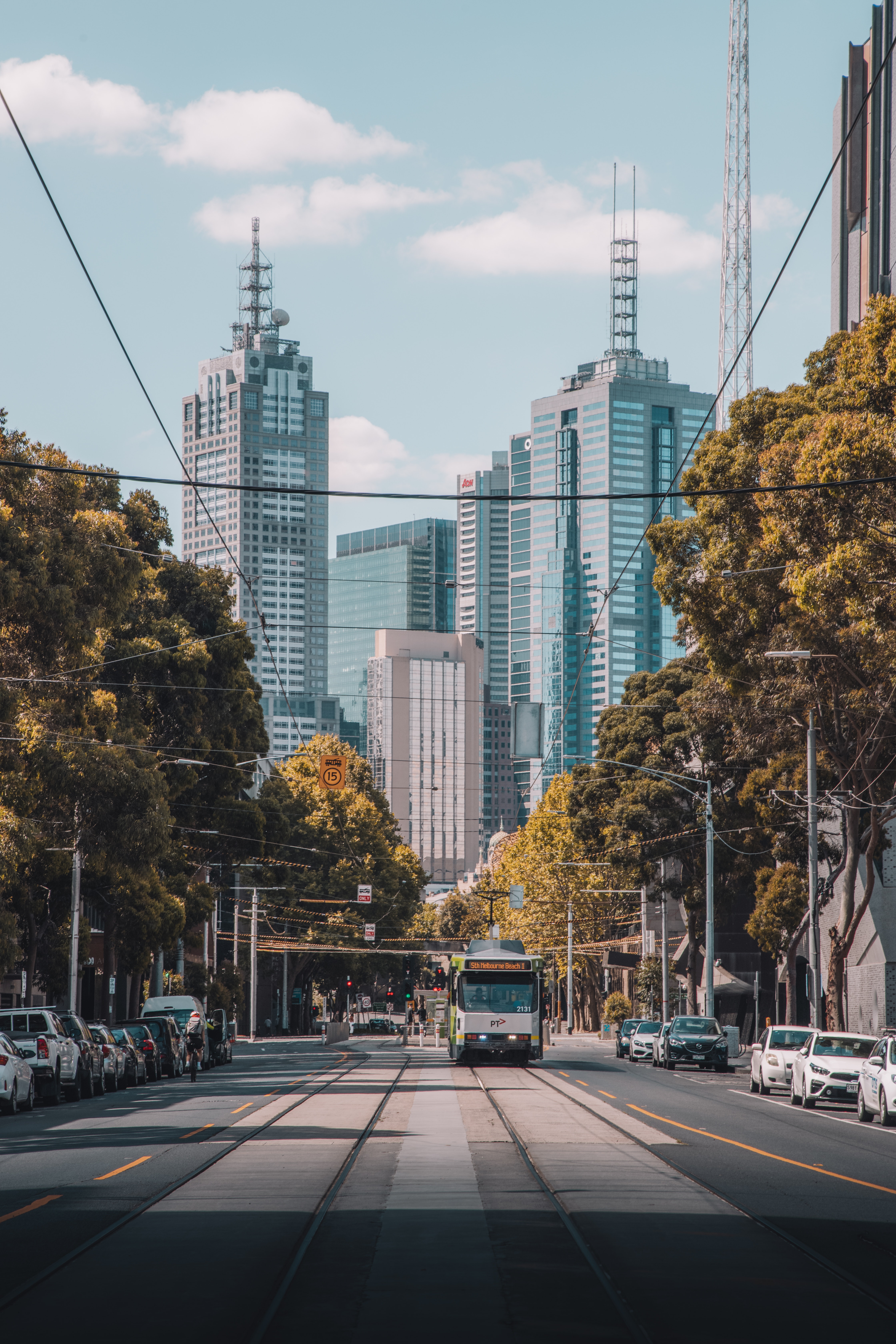 Image of Melbourne City Scape. The shot is taken with tram tracks at the forefront of the image with a tram in the centre of the composition. There are trees and cars on either side of the road and skyscrapers above. 