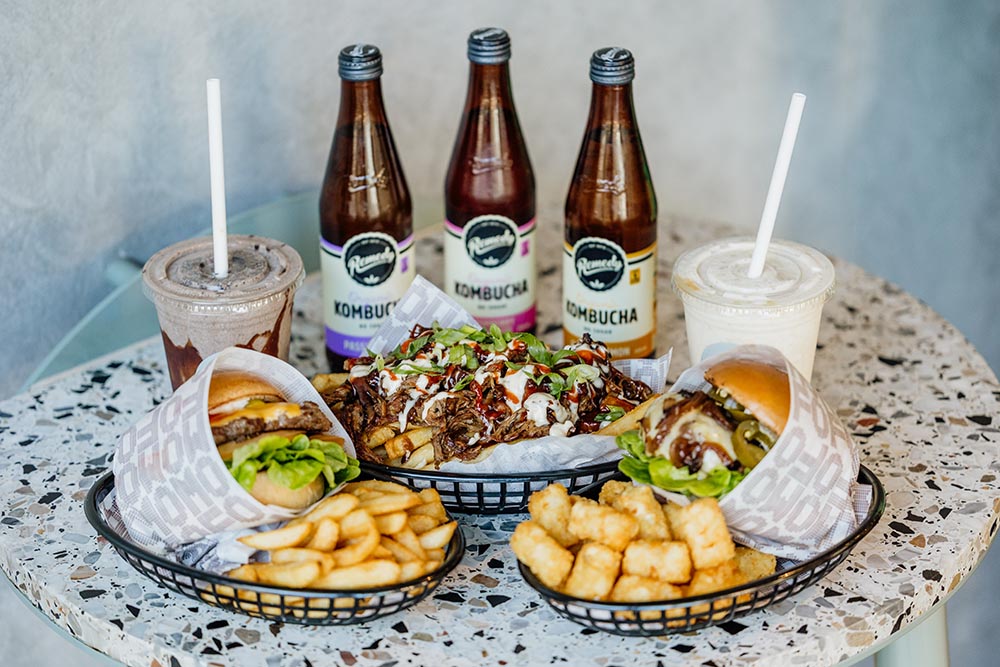 A table with two burgers, potato gems, an HSP, two shakes and three bottles of kombucha lined up