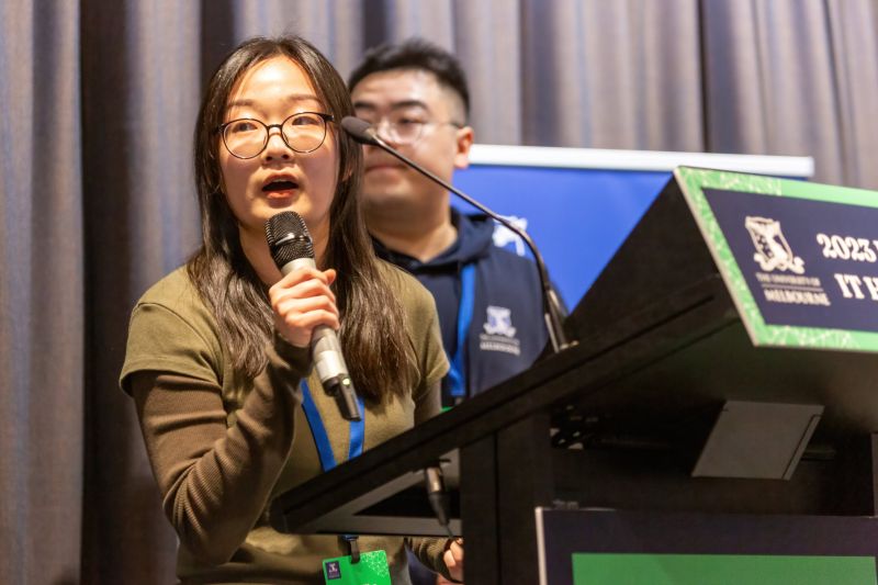 A woman stands in front of s alecturn speaking into a microphone. She has dark brown hair, glasses and a green shirt.