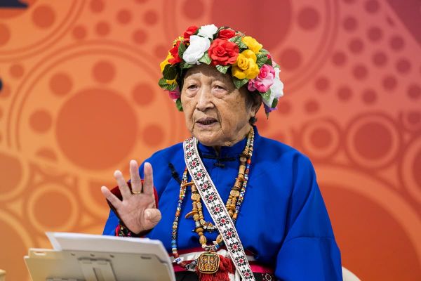Woman in floral headress, blue attire with various beads and adornments around her neck on an orange, patterned backdrop.