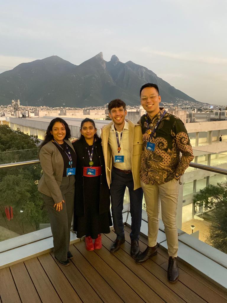 Four smiling students stand on a balcony with a large mountain and cityscape behind them at sunset.