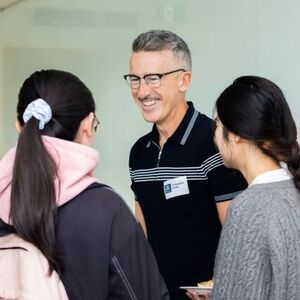 An alum wearing glasses and a polo shirt smiling while talking with two students.
                                class=
