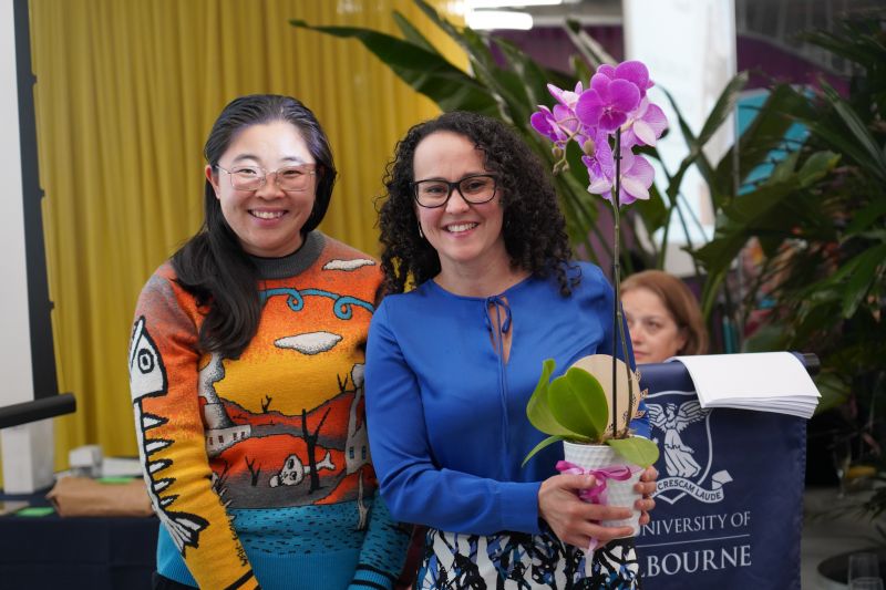 A dark haired woman in a yellow and blue jumper stands next to a dark haired woman in a blue top, carrying an orchid