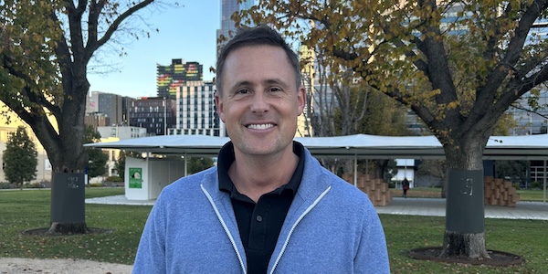 Headshot of Spencer dressed in a black shirt and blue jacket outside in a park