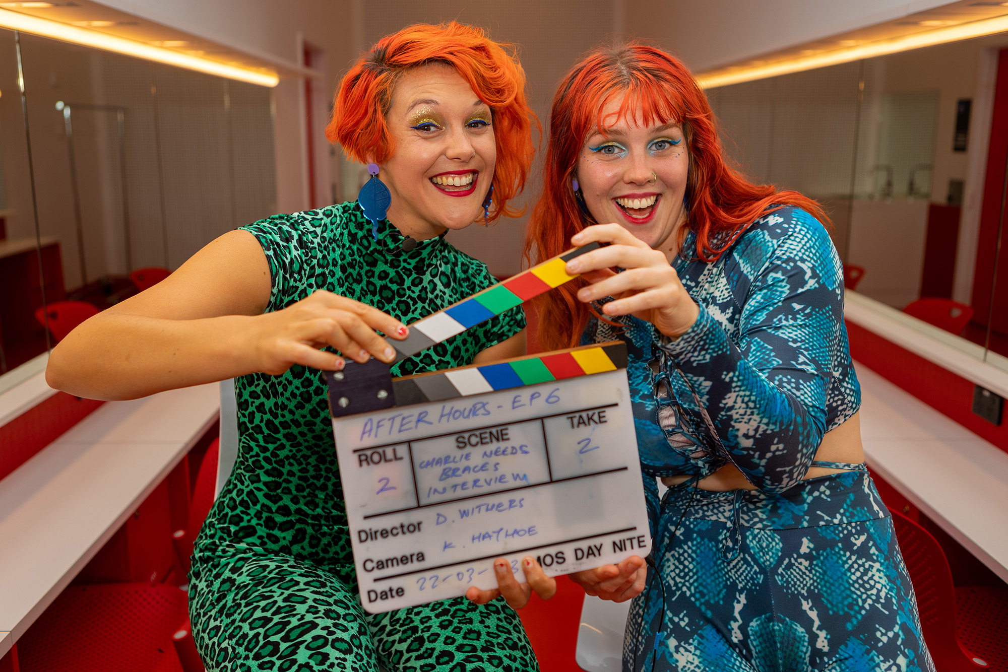 Two colourfully dressed women from Charlie Needs Braces pose with a clapperboard.