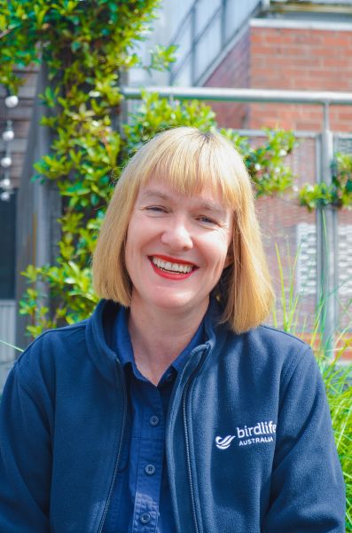 Person standing outdoors in front of greenery and a building, wearing a jacket with a visible &lsquo;BirdLife Australia&rsquo; logo