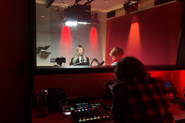 Podcast.jpg A studio operater looks through a glass window at a woman in a podcast studio.