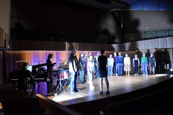 17 people stand on stage performing an opera, with various coloured lights and instruments playing in the background.