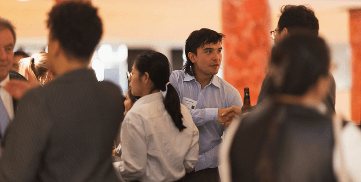 A group of alumni mingling at an event. The man in focus shakes hands with a fellow alum.