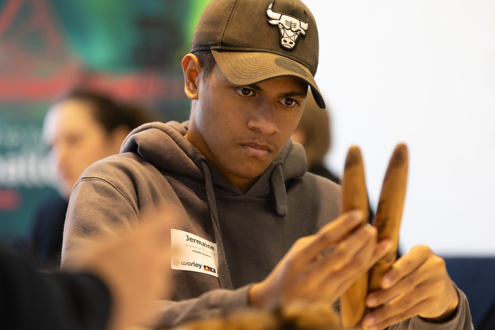 A young man in a hoodie and baseball cap holds a pair of wooden sticks, in a classroom setting.