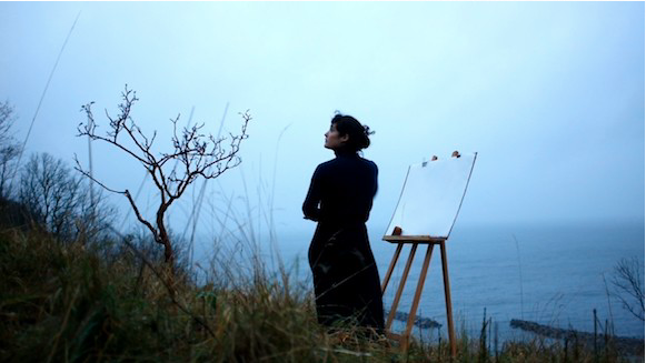 Video still depicting a woman on a hill side with a canvas on an easel in front of her.