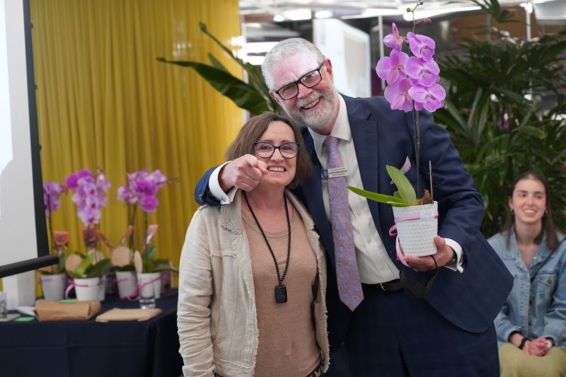 A man wearing a navy suit and carrying an orchid stands besides a woman with brown hair in a beige jacket an