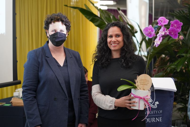 A woman with dark hair wearing and mask and wearing a blue suit stands next to a dark haired woman in black and white top holding an orchid