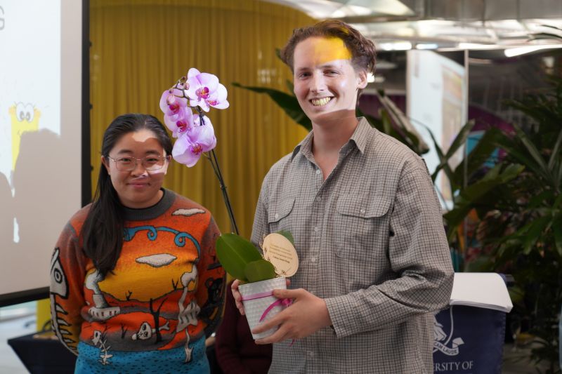 A dark haired woman in a colourful yellow and blue jumper stands next to a brown haired man in a grey shirt holding an orchid