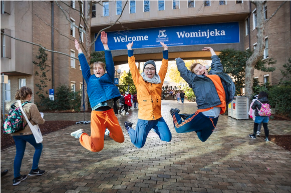 Image of students jumping in front of a large welcome banner at the main entrance of the University of Melbourne Parkville campus.