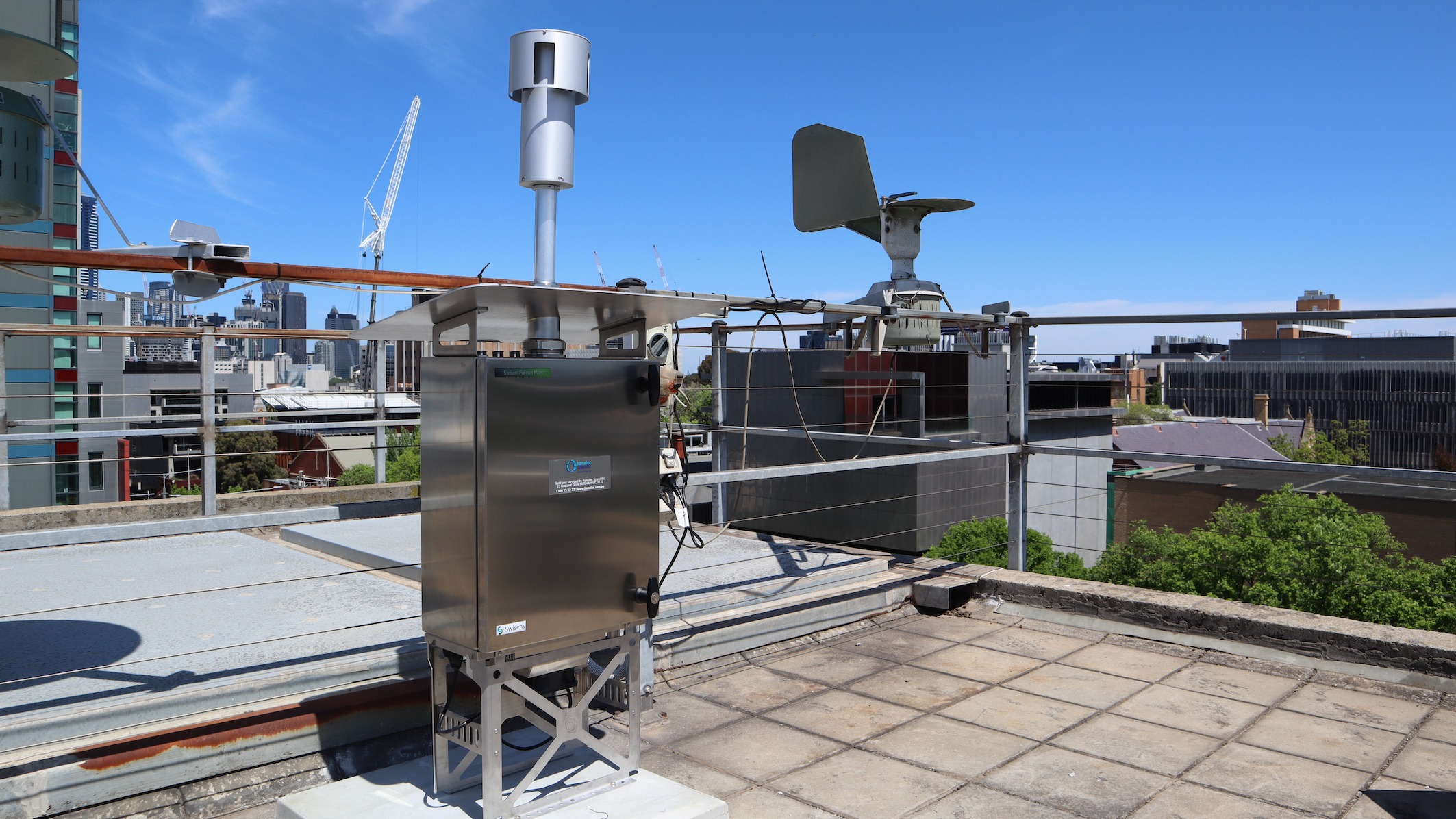 The pollen counter - a metal box with what looks like an antenna on top - sits atop a tall building from which you can see the blue sky and various other buildings on the University of Melbourne Parkville campus