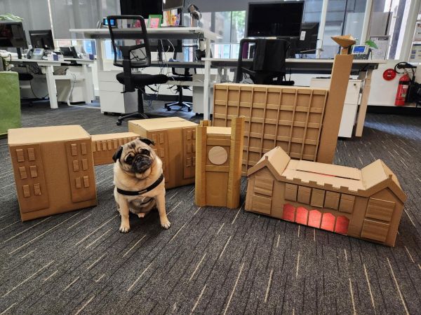 A small cute pug dog sits patiently in front of a cardboard cut out set of the University campus that was used in the Service Now videos.